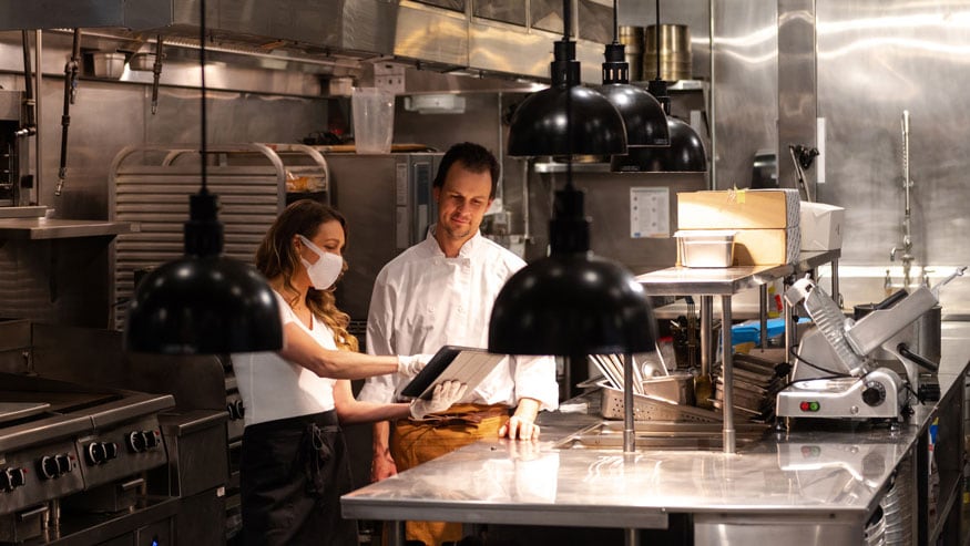 Two people in a restaurant kitchen. One is masked and pointing to the screen on a handheld device.