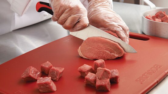 Kitchen staff holding a piece of meat on an Ecolab cutting board to be cubed with a commercial knife.