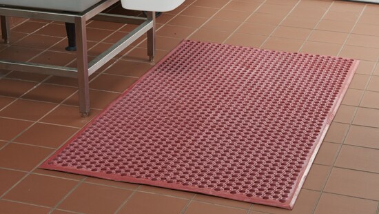 Ecolab red greaseproof floor safety mat laying on a tile floor next to a food prepping station in a commercial kitchen.
