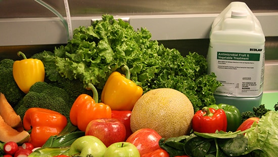 Cleaned fruits and vegetables on a cutting table next to a large bottle of antimicrobial fruit and vegetable treatment.