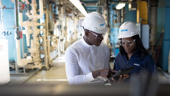 Two Ecolab experts in industrial room, filled with pipes and equipment, training.