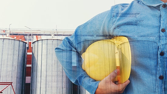 A person in a button-down blue shirt holding a yellow hard hat standing in front of a building. 