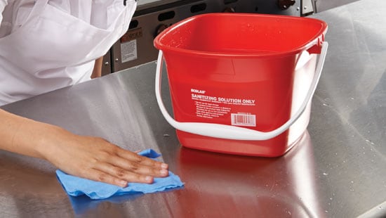 Employee cleaning prep table in a commercial kitchen with a dish cloth and bucket filled with sanitization solution.