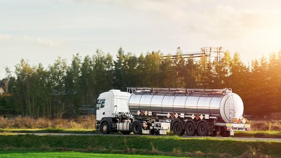 Stainless steel fuel tanker truck driving on a rural road with trees and sunlight in the background.