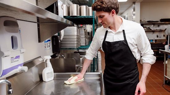 Person cleaning a stainless-steel counter in a commercial kitchen using a cloth and cleaning solution.