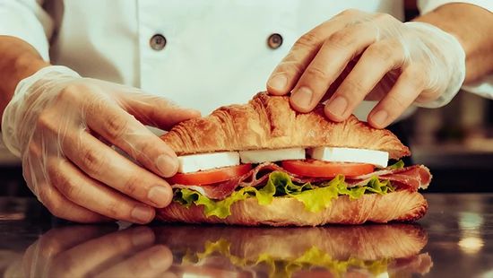 Gloved hands prepare a fresh croissant sandwich with deli meat, cheese, lettuce and tomato.