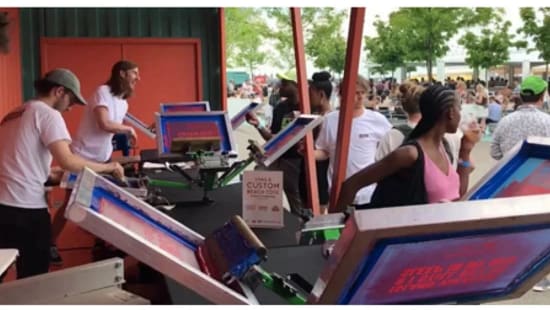 Outdoor event booth with multiple screen‑printing stations where staff are printing custom designs on apparel while attendees wait nearby.