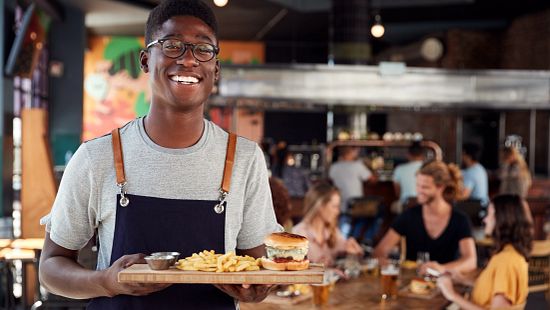 A restaurant staff member holding a tray with a burger, fries, and a sauce cup, standing in a casual dining space with seated guests in the background.
