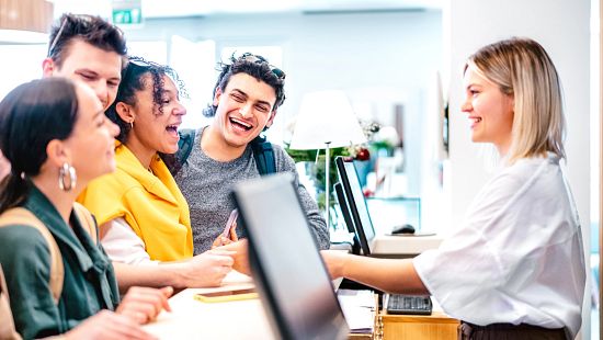 Group of young adults smiling and chatting with a staff member at a counter in a bright indoor space.