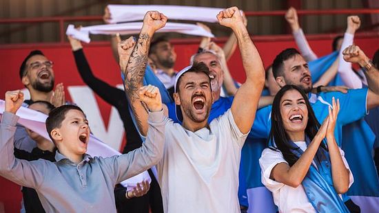 A group of people in stadium seating raise their arms and clap together, with several holding fabric banners or flags, suggesting a live sporting or public event in an indoor arena.