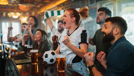 A group of people gathered around a bar counter with drinks and a soccer ball, watching a sports match on screens inside a pub.