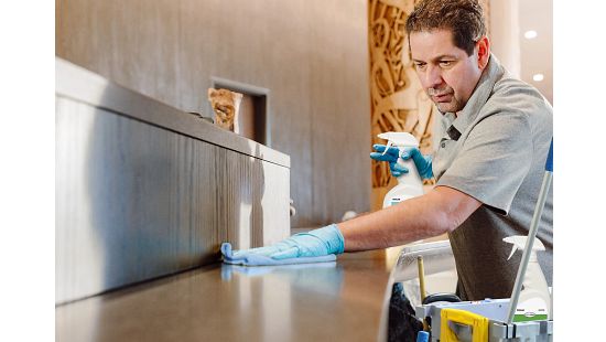 Person wearing gloves cleaning a countertop with a spray bottle and cloth, with a cleaning cart nearby.