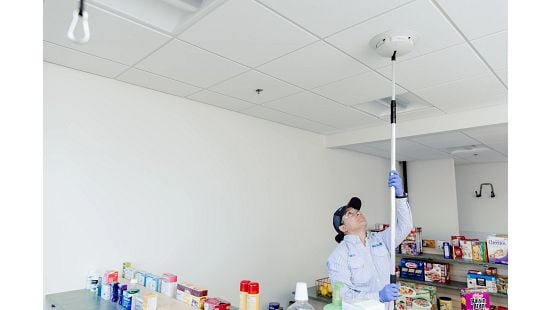 Person in uniform using a pole to adjust a ceiling device in a pantry with food shelves.